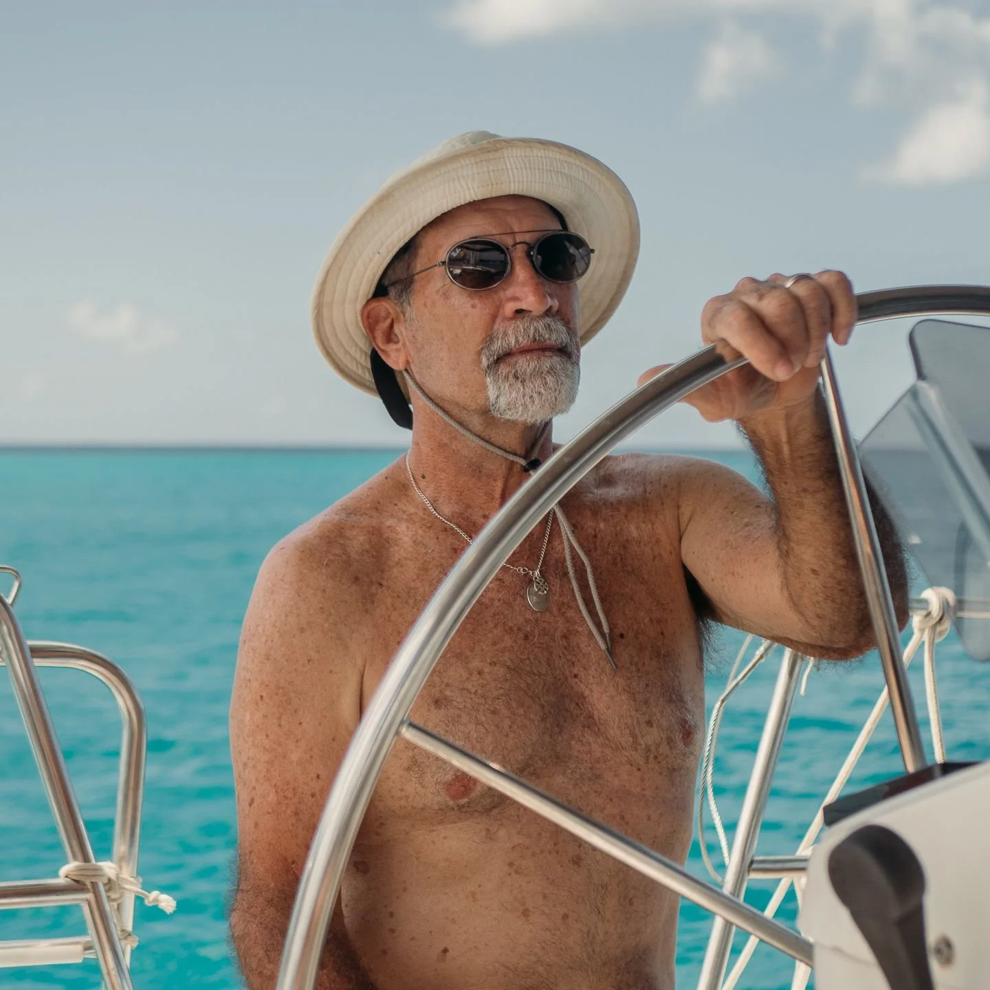 A man wearing a hat and sunglasses at the helm of Nayru, sailing on the coast of Barbados on a sunny day.