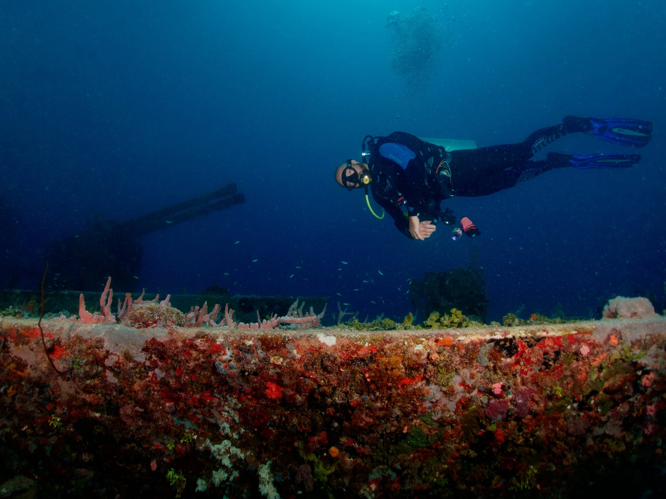 A scuba diver swimming along the deck of the shipwreck of the MV Trident in Carlisle Bay, Barbados with the replica guns in the background.