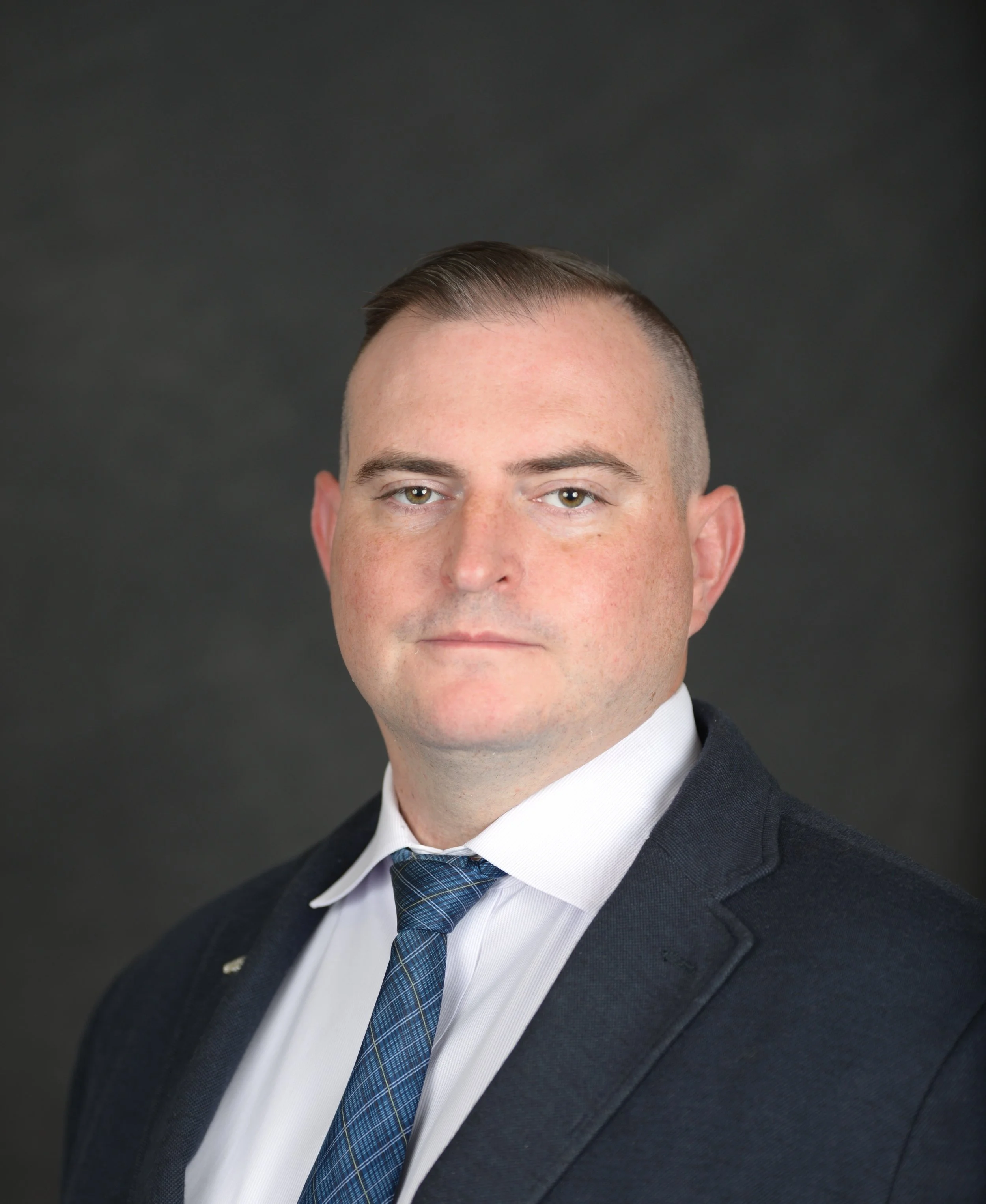 Professional headshot of a man in a dark suit, white shirt, and blue plaid tie against a dark background.