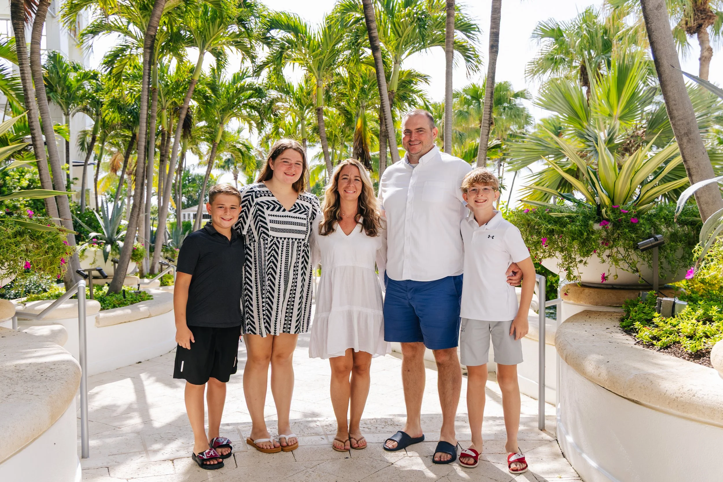A family of six standing together outdoors among palm trees and tropical plants, smiling at the camera.