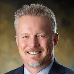 Portrait of a smiling middle-aged man with short gray hair, dressed in a suit, against a blurred yellow and black background.
