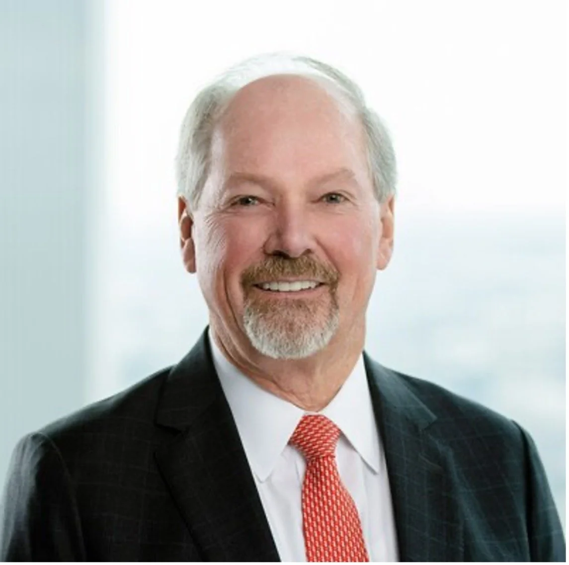 A smiling middle-aged man with gray hair and a beard, wearing a dark suit with a white shirt and red tie, posing in front of a blurred interior background.