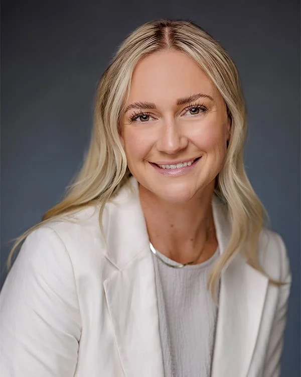 Portrait of a smiling woman with blonde hair, wearing a white blazer and a light-colored top against a dark background.