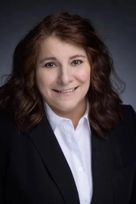 Professional woman with shoulder-length brown hair wearing a navy blazer and white shirt, smiling at the camera against a dark background.