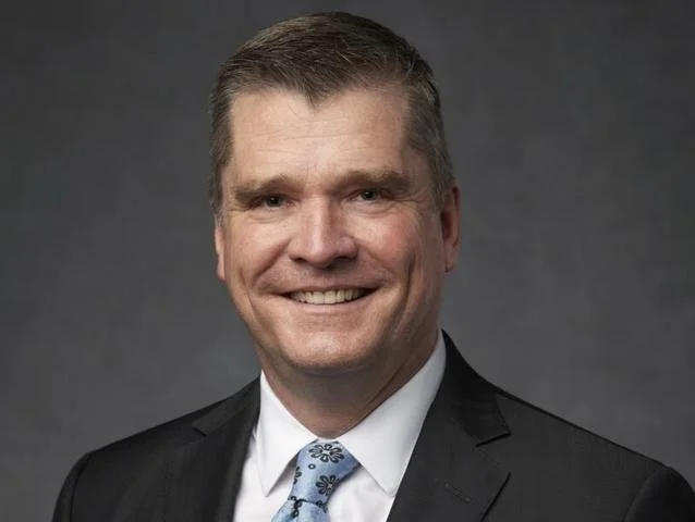 Headshot of a smiling man in a suit with a dark gray background.