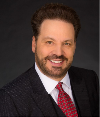 Headshot of a man with dark hair, beard, wearing a suit and red tie, smiling against a dark background.