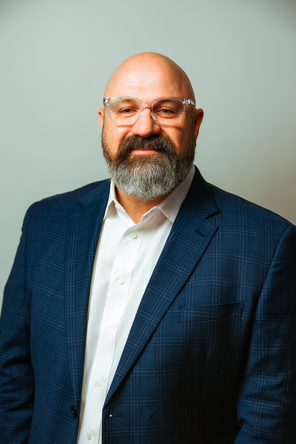 A middle-aged man with a beard and glasses, dressed in a white dress shirt and a dark blue plaid suit, posing against a plain light-colored wall.