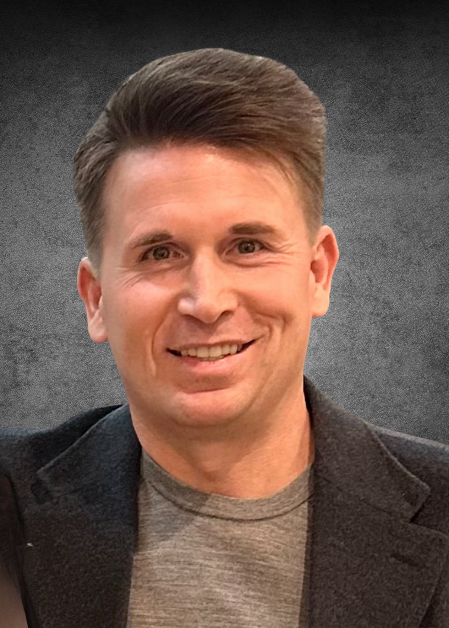 Smiling man with brown hair wearing a gray T-shirt and a dark blazer, posing in front of a gray textured background.
