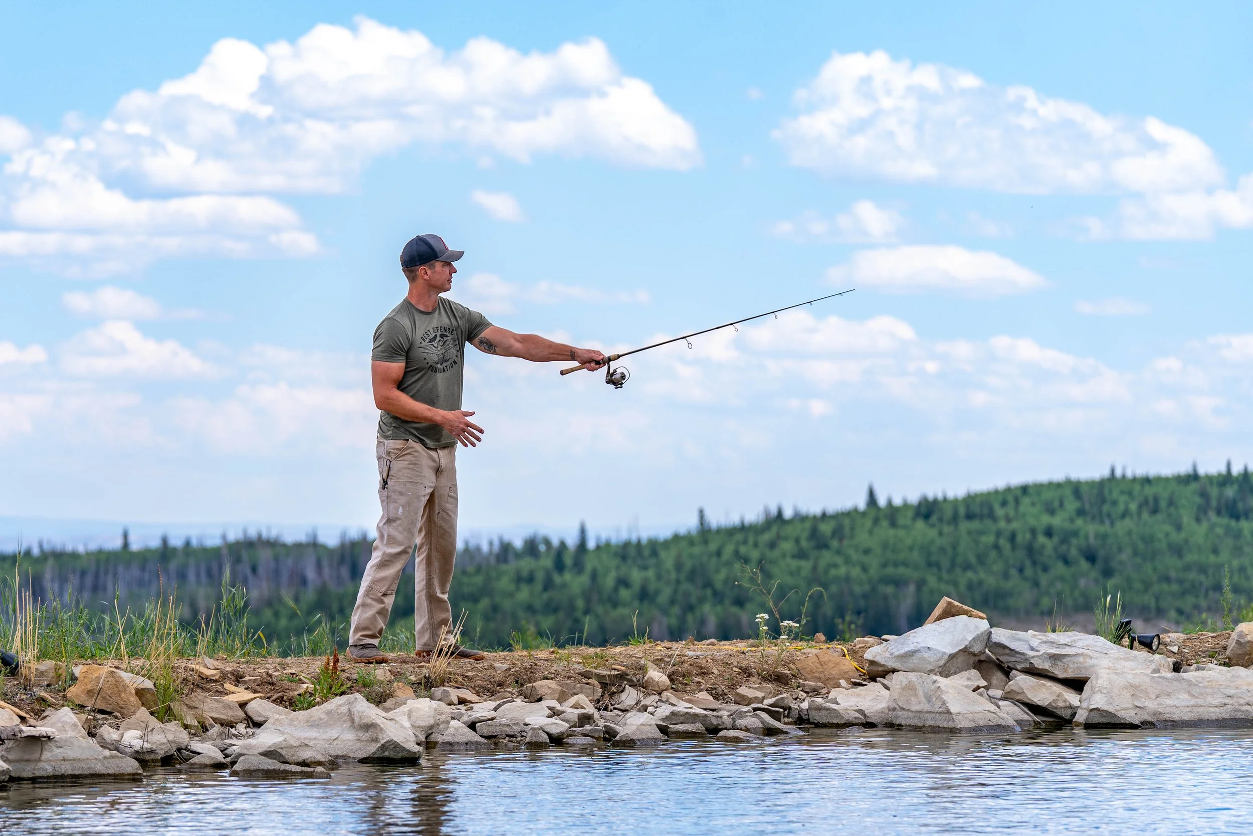 A man standing on rocky shore fishing in a river or lake with a fishing rod, wearing a cap, t-shirt, and khaki pants, against a background of green hills and blue sky with clouds.