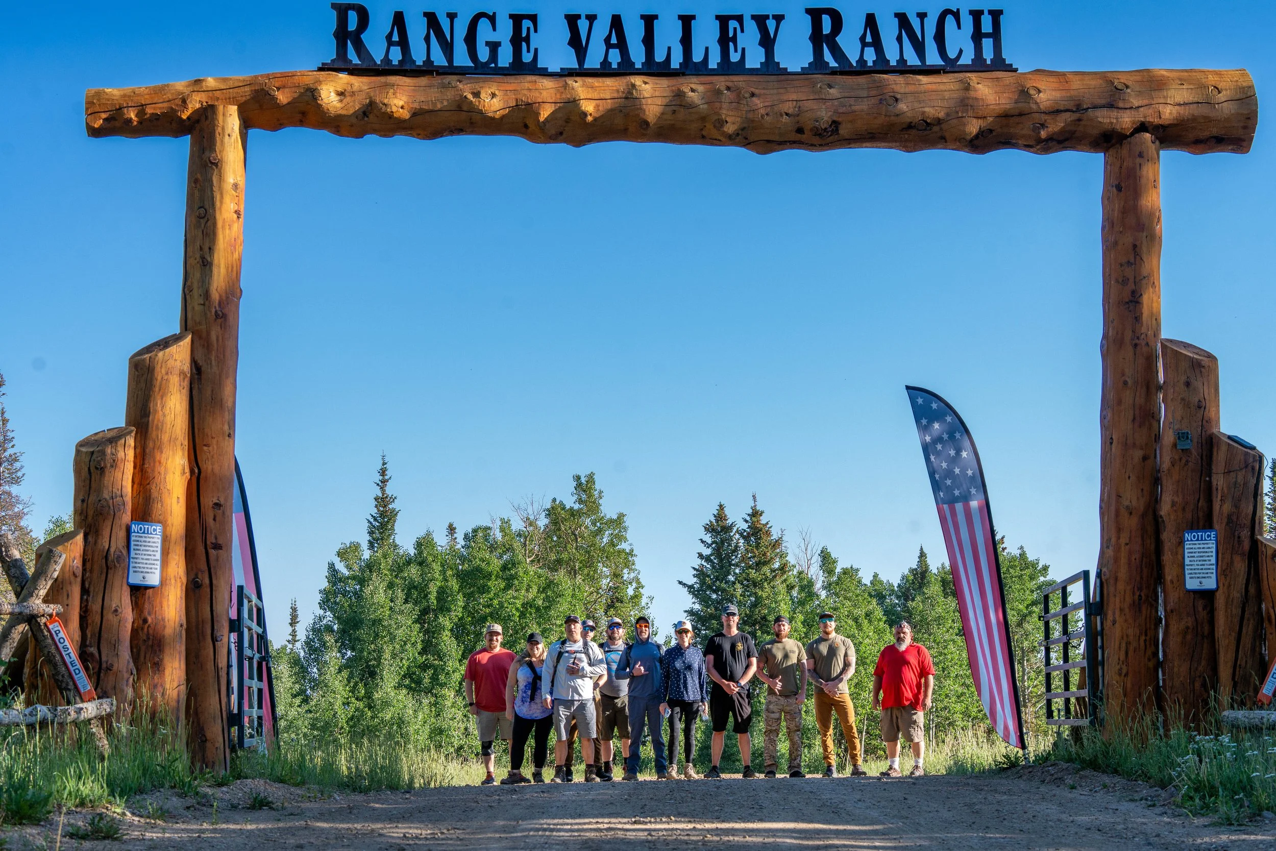 A group of people standing under a large wooden archway with the sign 'Range Valley Ranch' at the top, surrounded by trees and a clear blue sky, with American flags on either side of the arch.