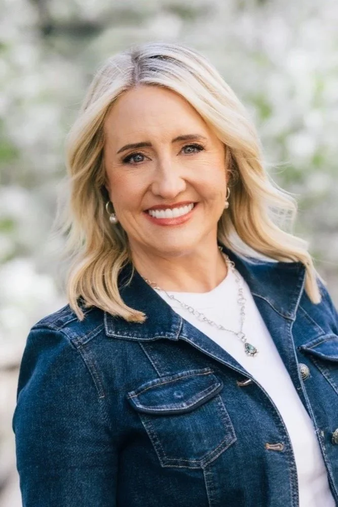 A smiling blonde woman with wavy hair, wearing a denim jacket, white shirt, and jewelry, outdoors with blurred greenery in the background.