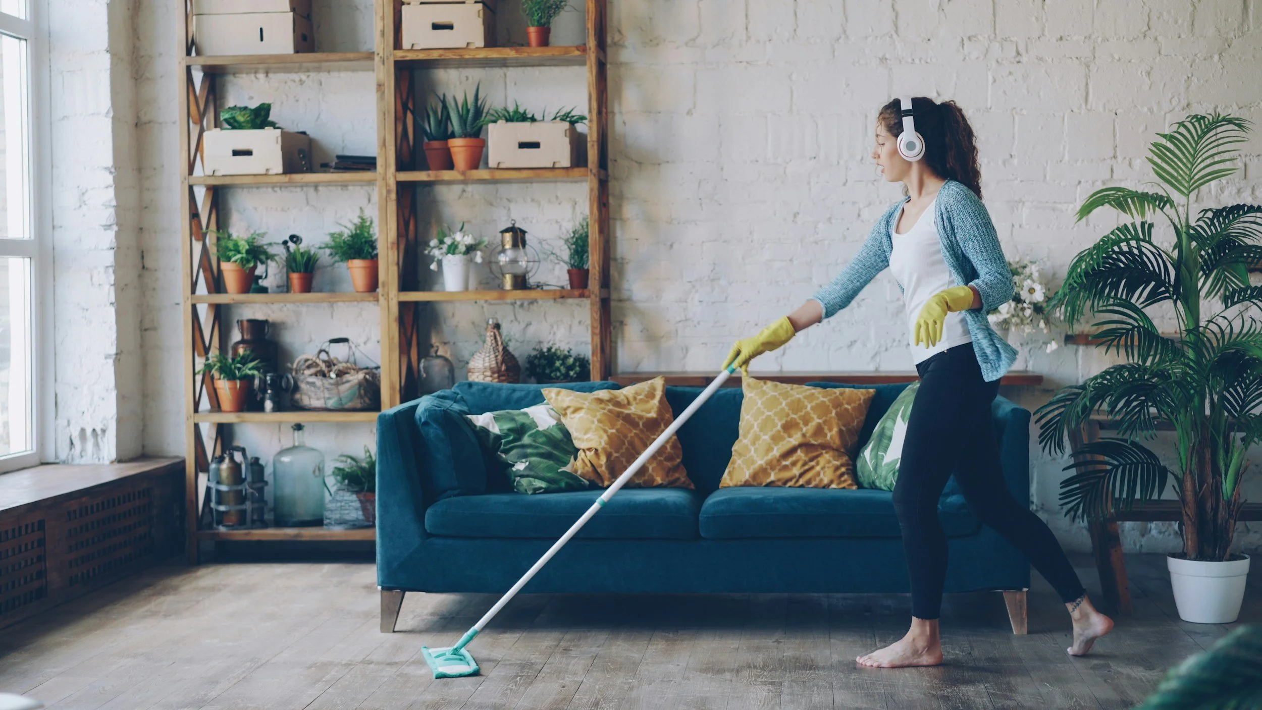 Woman cleaning the floor with a mop in a living room with white brick walls, wooden shelf with plants and decor, a blue sofa with yellow and green pillows, and large potted plants.