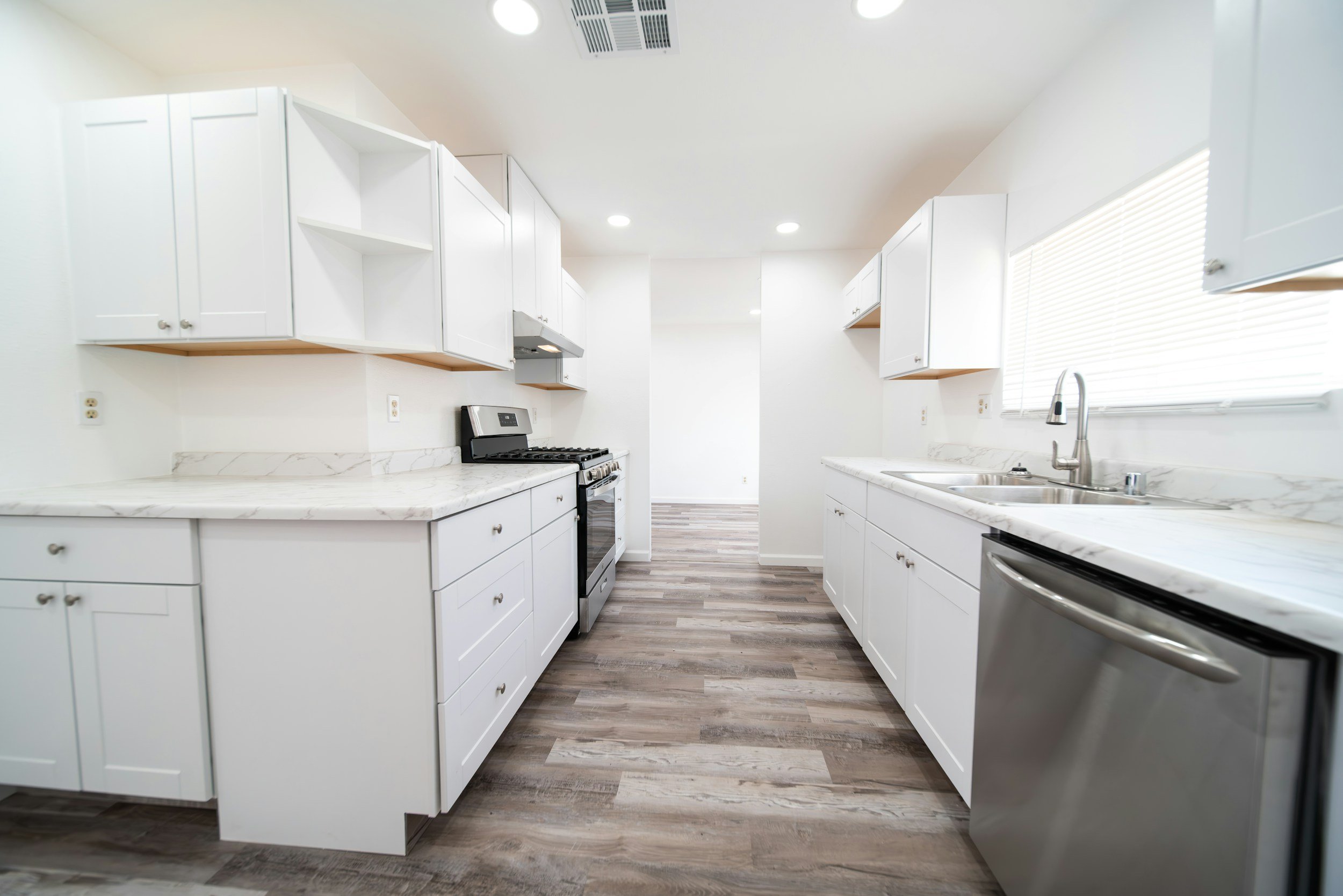 A modern kitchen with white cabinets, marble countertops, and stainless steel appliances. There is a double sink under a window with closed blinds, and a stove in the middle of the left counter. The floor has wood-look laminate flooring.