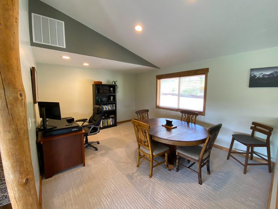 Home office and dining area with a computer desk and chair, a wooden dining table with four chairs, a window with wooden blinds, a bookshelf, and a framed picture on the wall.