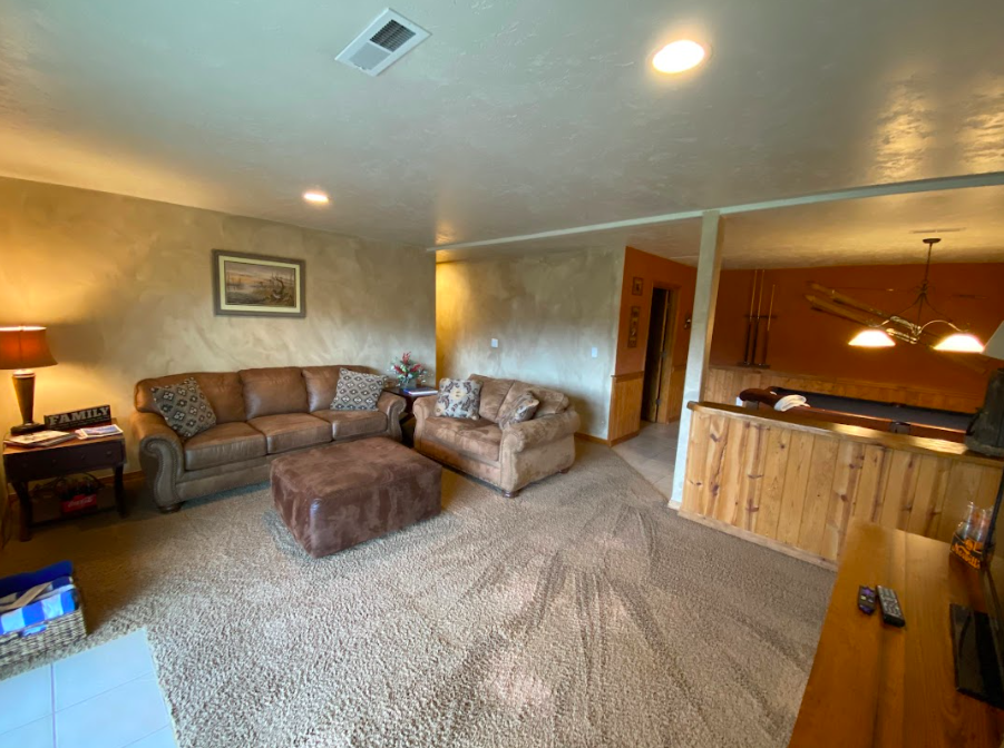 Living room with a brown couch, a matching armchair, and an ottoman, with a side table and a lamp on the left. There is a framed picture on the wall and a small flower arrangement. The area is carpeted with neutral-colored carpet, and part of a dinin