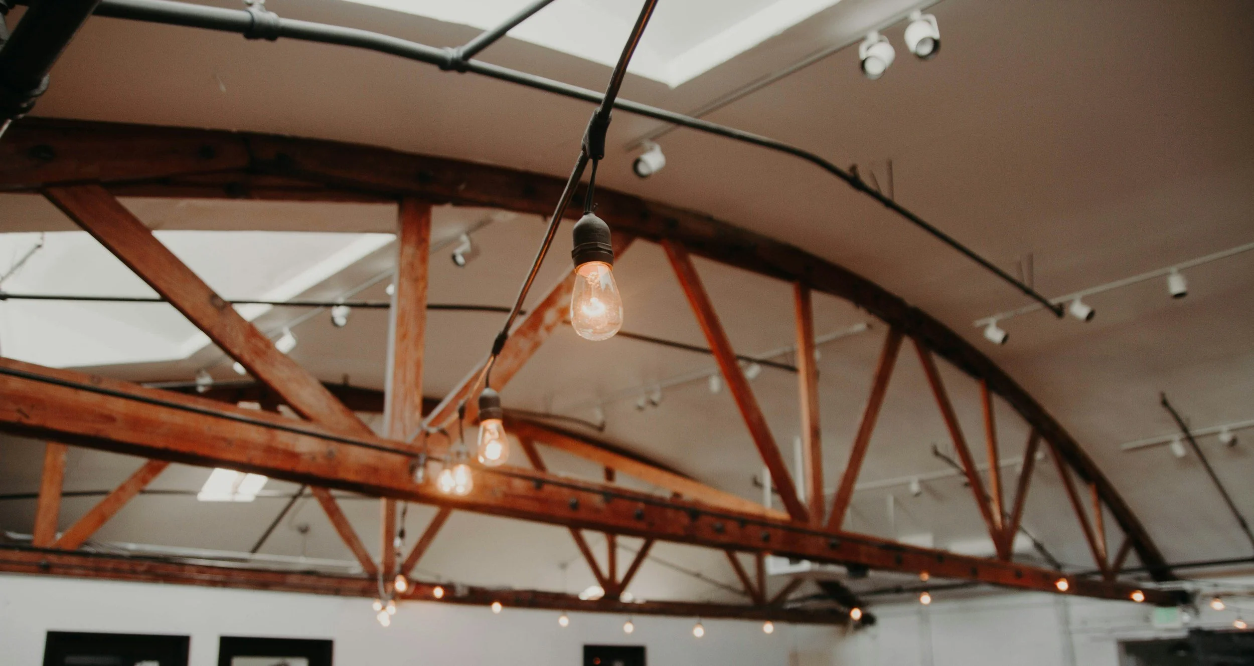 Interior of a room with a curved ceiling, exposed wooden beams, and string lights hanging, with some pendant lights turned on.