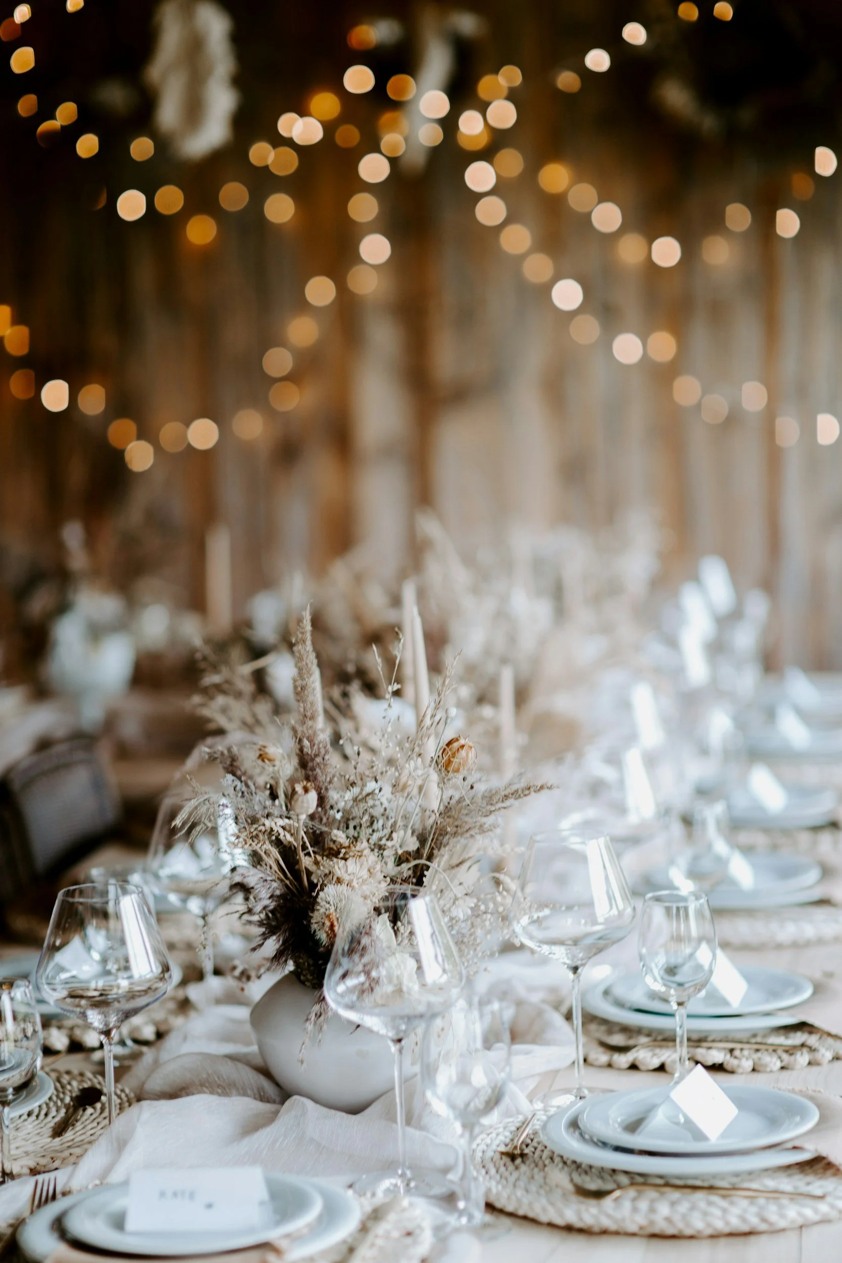 A beautifully set rustic wedding reception table with a floral centerpiece, white plates, and glassware under string lights in a wooden barn.