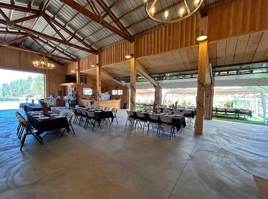 Interior of a rustic event barn with tables set for a gathering, featuring black tablecloths, wooden accents, and outdoor views through open sections.