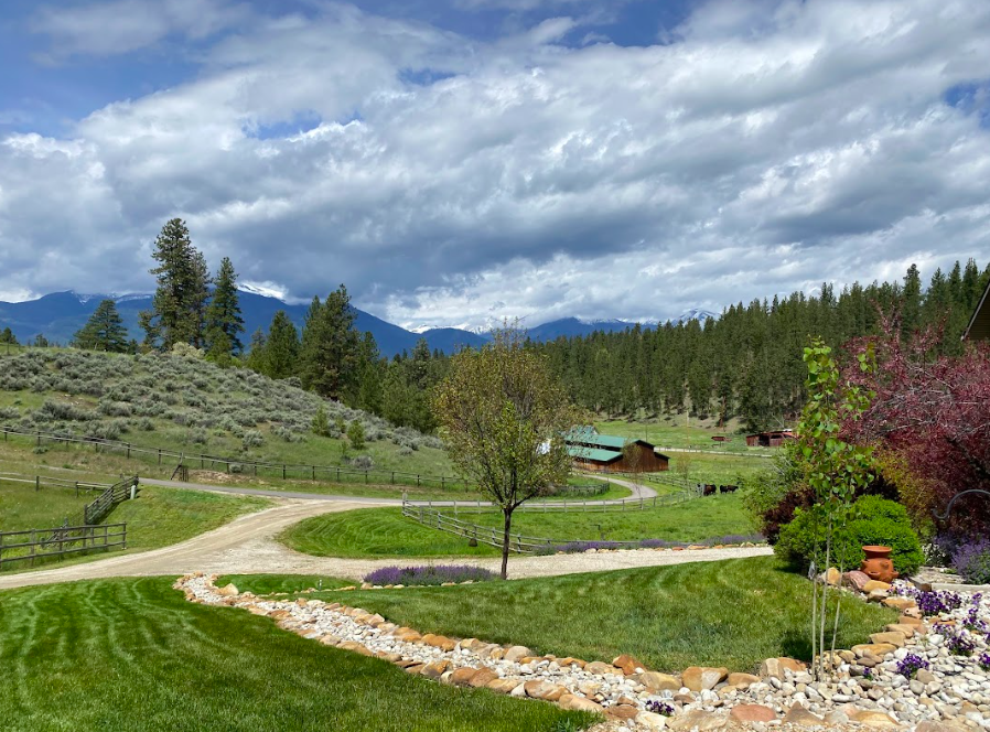A scenic landscape with a lush green lawn, a winding gravel driveway, trees, a rustic barn, and a mountain range with snow-capped peaks in the background under a cloudy sky.