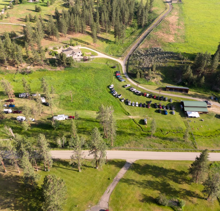 A rural area with a few buildings, a parking lot filled with cars, a road, and green grassy fields surrounded by trees.