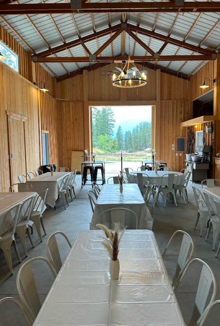 Interior of a rustic wooden event space with tables covered in white tablecloths, white chairs, and a view of trees and mountains through large window doors at the back.