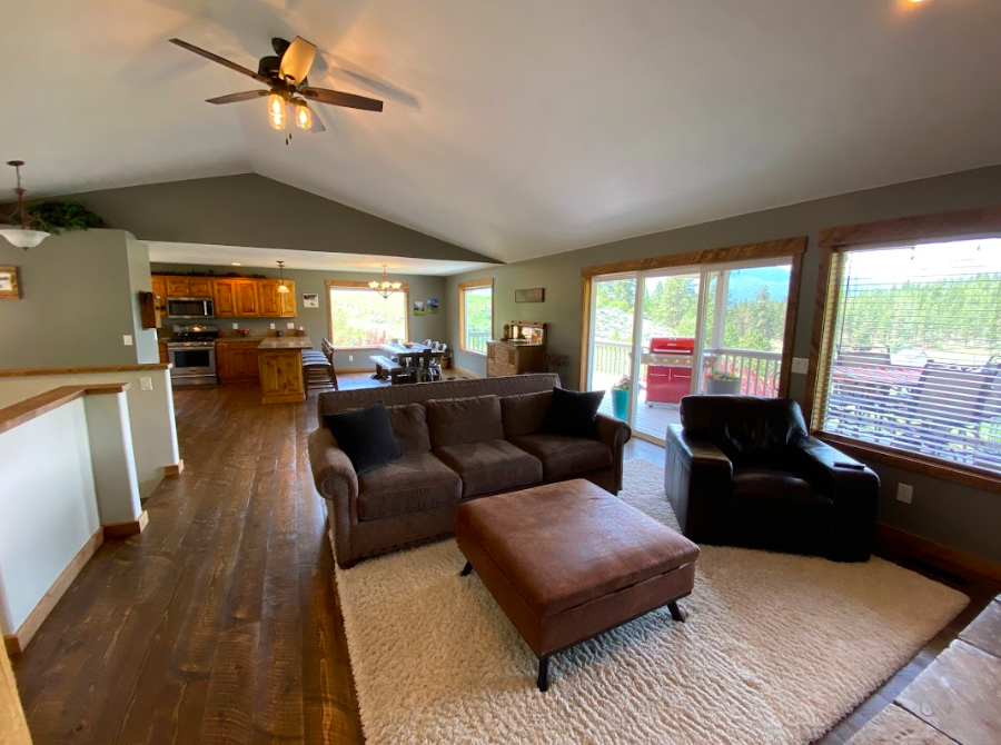 Living room with a brown sofa, black armchair, and ottoman on a beige rug, large windows and sliding glass door leading to a deck, view of trees outside, kitchen area in the background with wooden cabinets and dining table.