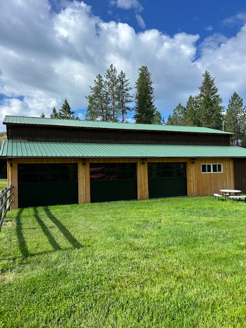 A wooden building with a green metal roof, three large garage doors, and a small window, set in a grassy area with trees in the background and a partly cloudy sky.
