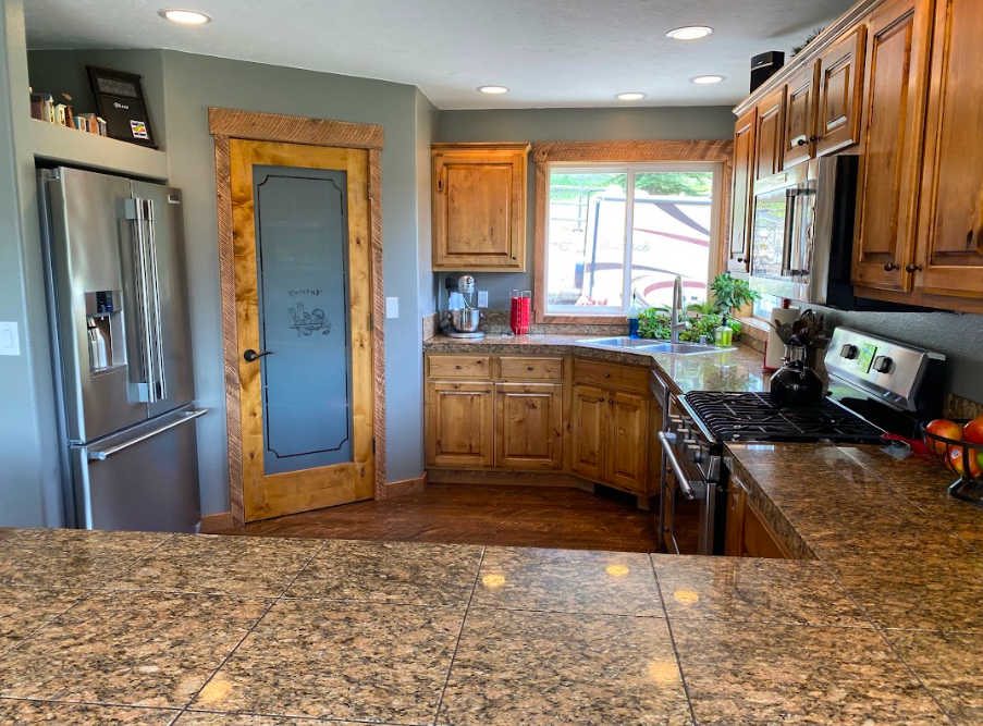 A rustic kitchen with wooden cabinets, granite countertops, a stainless steel refrigerator, and a window above the sink with greenery outside.