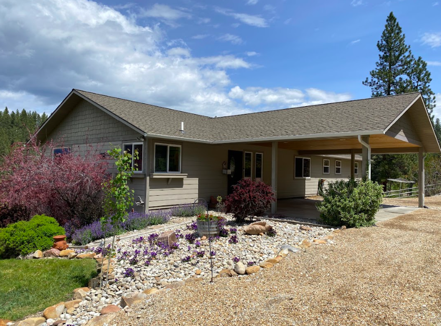 Residential house with landscaped front yard, including bushes, flowers, and a gravel pathway, under partly cloudy sky.