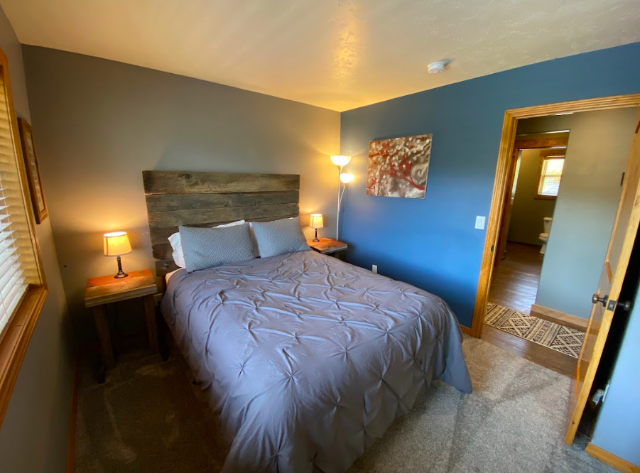 A cozy bedroom with a wooden headboard, gray bedding, and two bedside tables with lamps. There is a window with blinds on the left, a blue accent wall, and a framed floral painting. An open door shows a bathroom with a toilet and a small window.