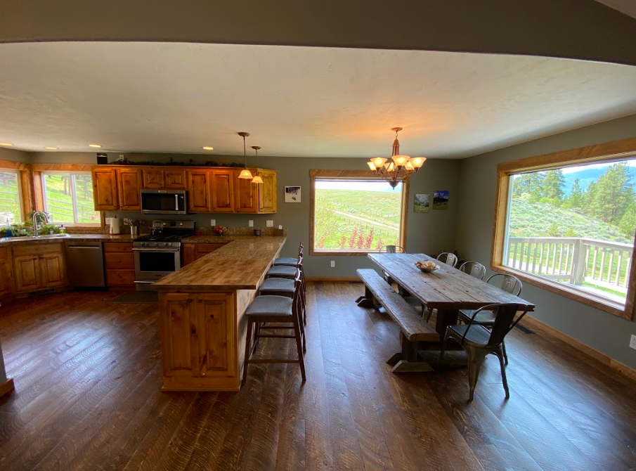 Open kitchen and dining area with large windows showcasing green landscape, wooden furniture, and a chandelier.