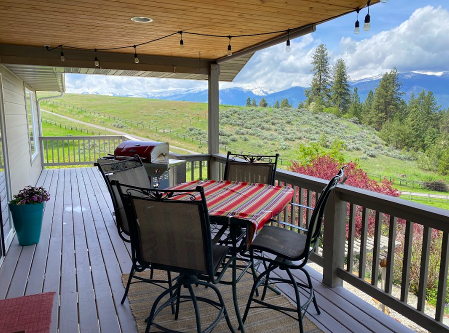 Outdoor porch with a table covered with a striped tablecloth, six chairs, a red grill, a turquoise pot with flowers, overlooking a green landscape with rolling hills, trees, and mountains in the distance under a partly cloudy sky.