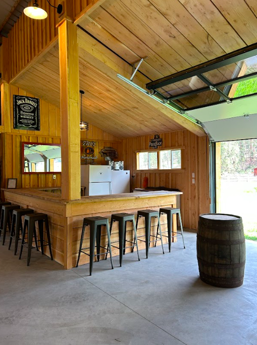 Indoor bar area with wooden walls and ceiling, black bar stools, a barrel, and an open garage door showing greenery outside.