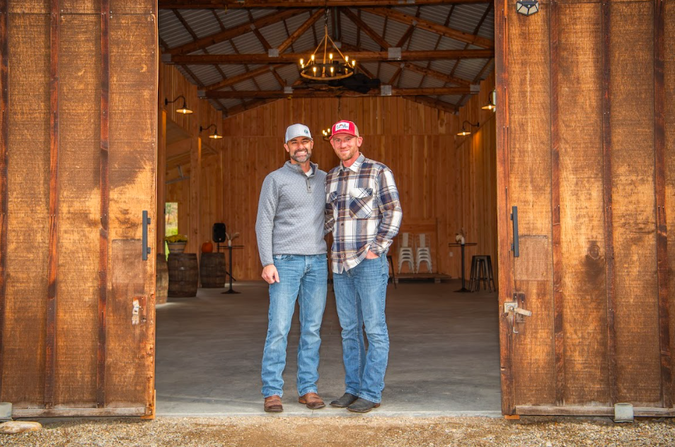 Two men standing inside a rustic wooden barn, smiling at the camera. One is wearing a gray cap and gray sweater, and the other is wearing a red cap and plaid shirt. The barn has wooden walls, a chandelier above, and a few chairs in the background.