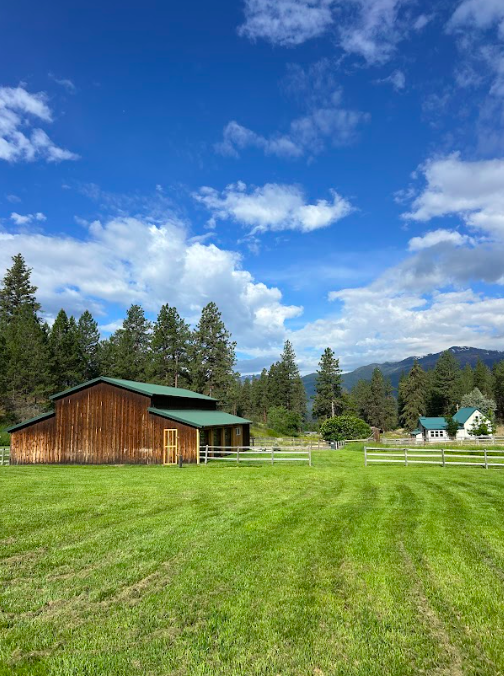 A rural scene with a brown wooden barn, a white fence, green grass, trees, and a partly cloudy sky with mountains in the background.