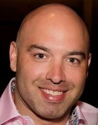 A smiling man with short reddish-brown hair wearing a plaid shirt