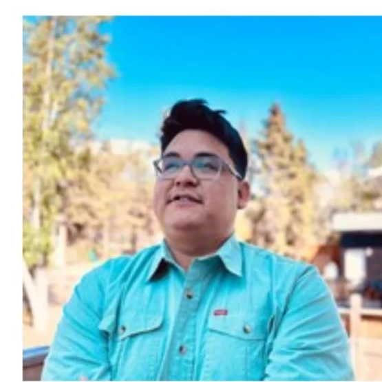 Johnny Ongahak, a smiling man wearing glasses and a teal button-up shirt outdoors.
