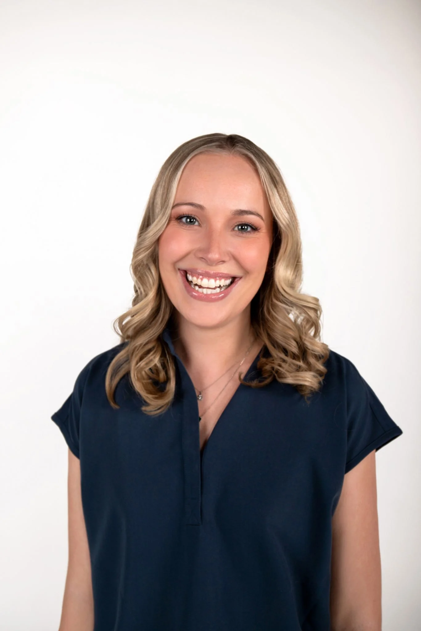 A young woman with blonde curly hair, smiling, wearing a navy blue top, standing against a plain white background.