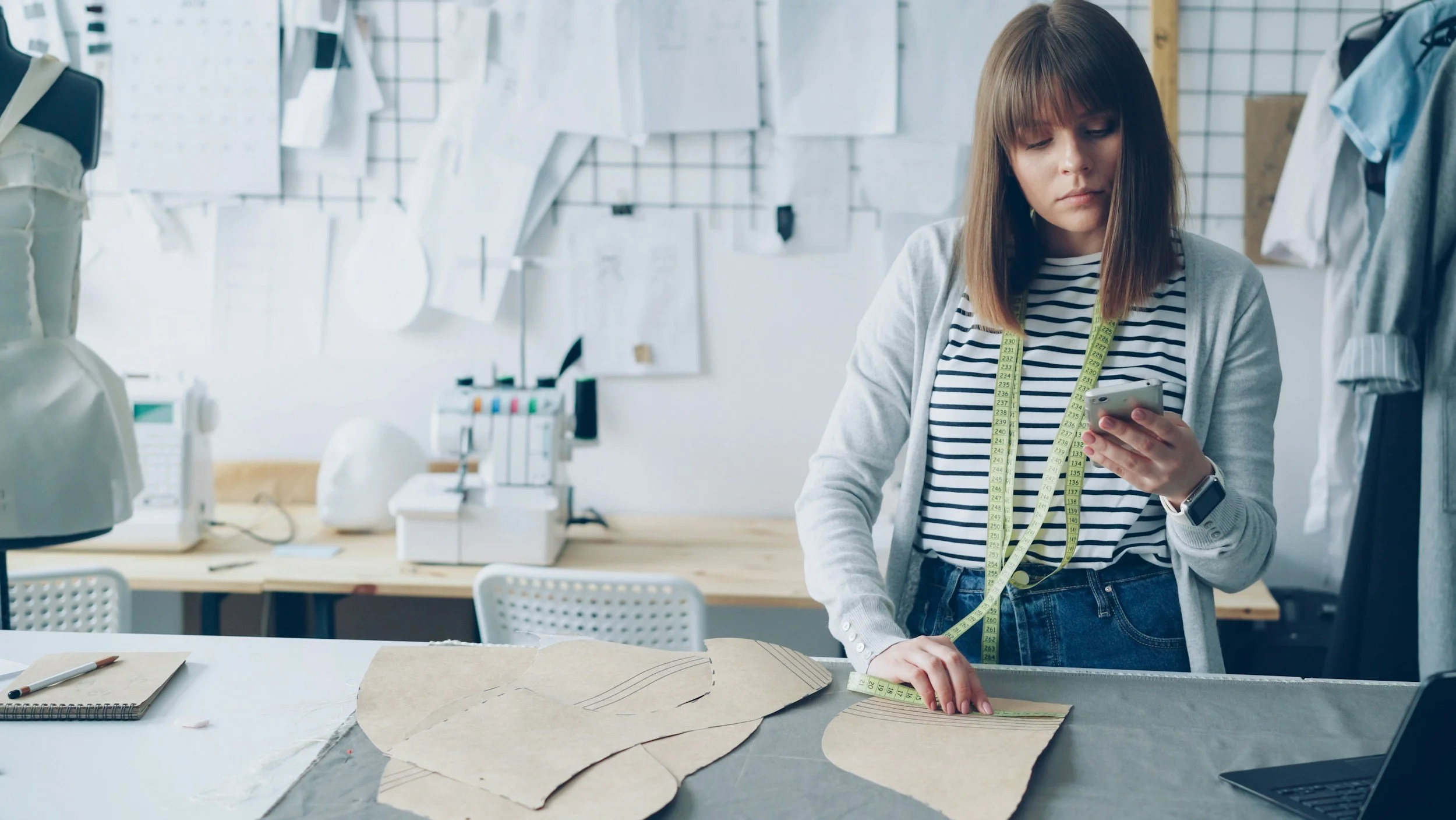 A young woman with a measuring tape around her neck is working in a fashion design studio. She is holding her phone in one hand and measuring fabric with the other. There are sketches and fabric pieces on the table in front of her, and a sewing machine and dress form in the background.
