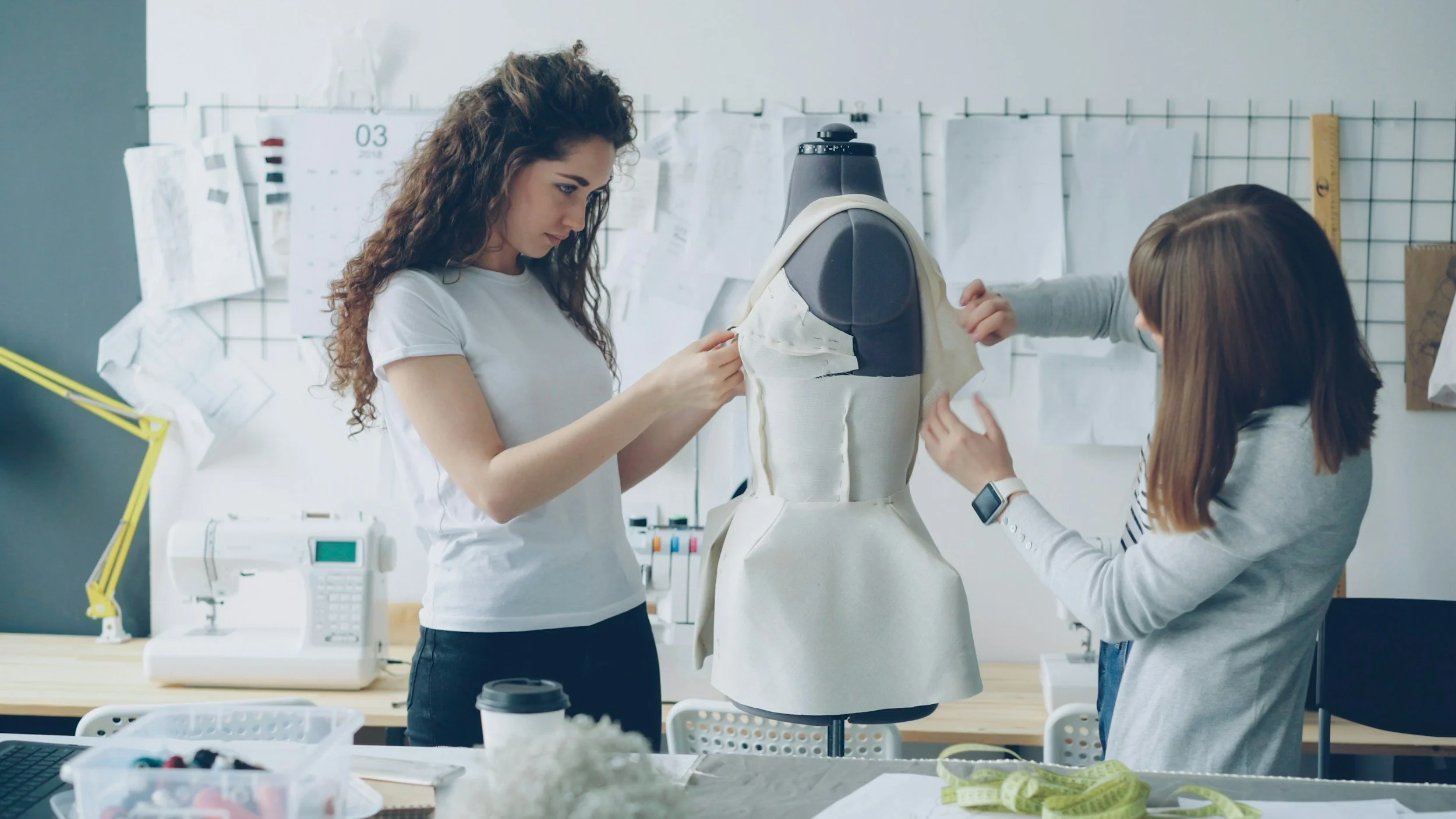 Two women working on a fashion design dress form in a design studio, surrounded by sewing and design tools.
