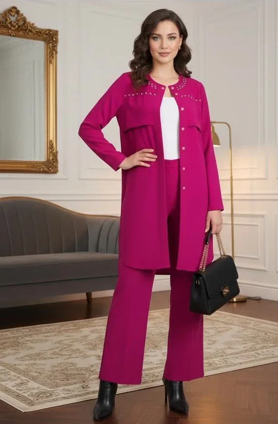Woman standing in a room wearing a vibrant pink coat, matching pink pants, black high-heeled boots, and holding a black handbag with a gold chain, next to a gray couch and a gold-framed mirror.