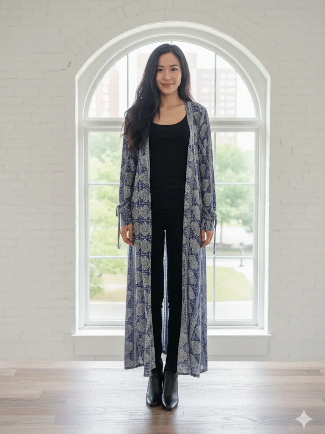 A woman with long dark hair standing indoors in front of a large arched window, wearing a black top, patterned long cardigan, black pants, and black boots.