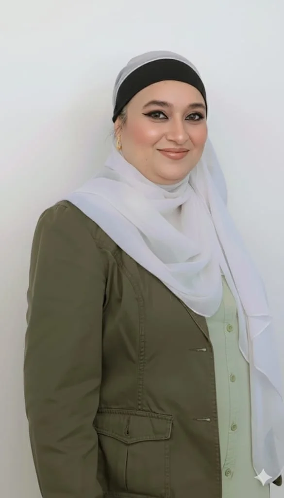 A woman wearing a white hijab, an olive green jacket, and gold earrings, smiling and standing against a plain white background.