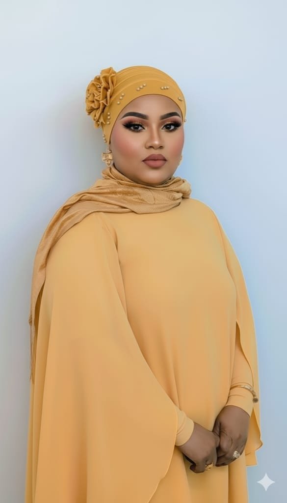 A woman wearing a beige hijab with floral details and matching gown, standing against a plain light blue background.