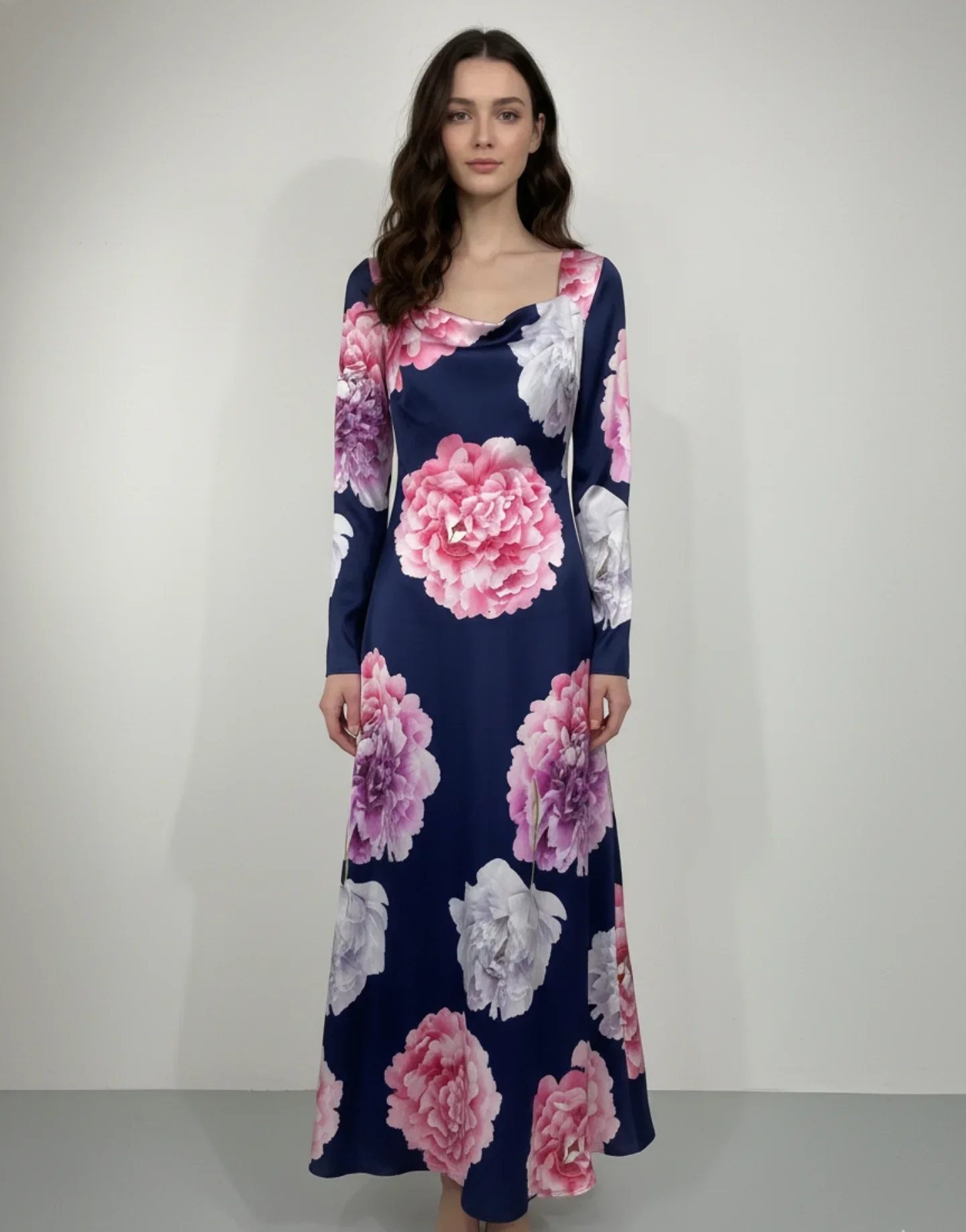 A woman with long wavy brown hair wearing a long, dark blue floral dress with pink and white flowers standing against a plain white wall.