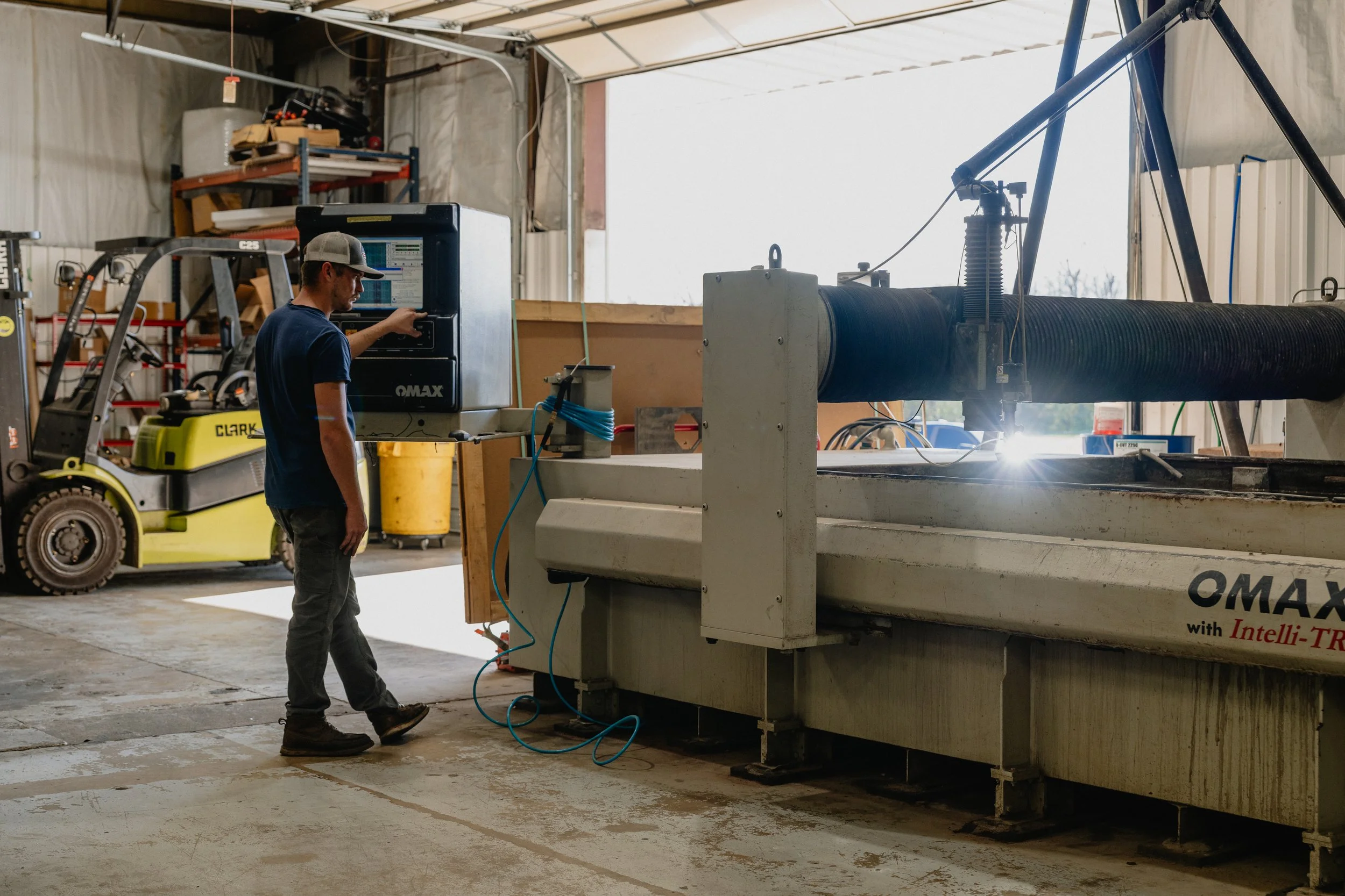 A person operating a CNC machine inside a workshop, with a forklift and other equipment visible in the background.