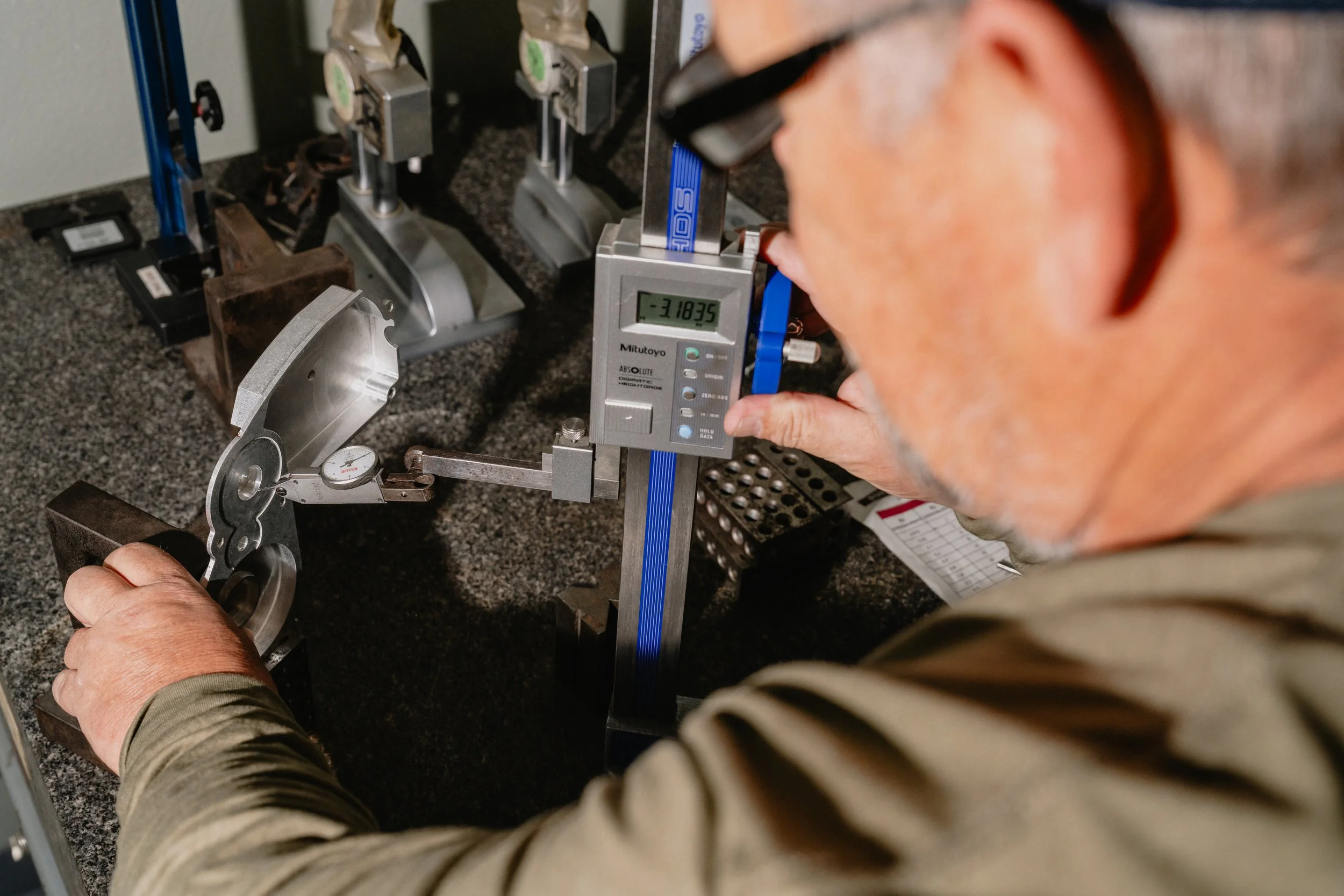 A person measuring a metal part with a digital caliper on a workbench containing various mechanical tools and equipment.