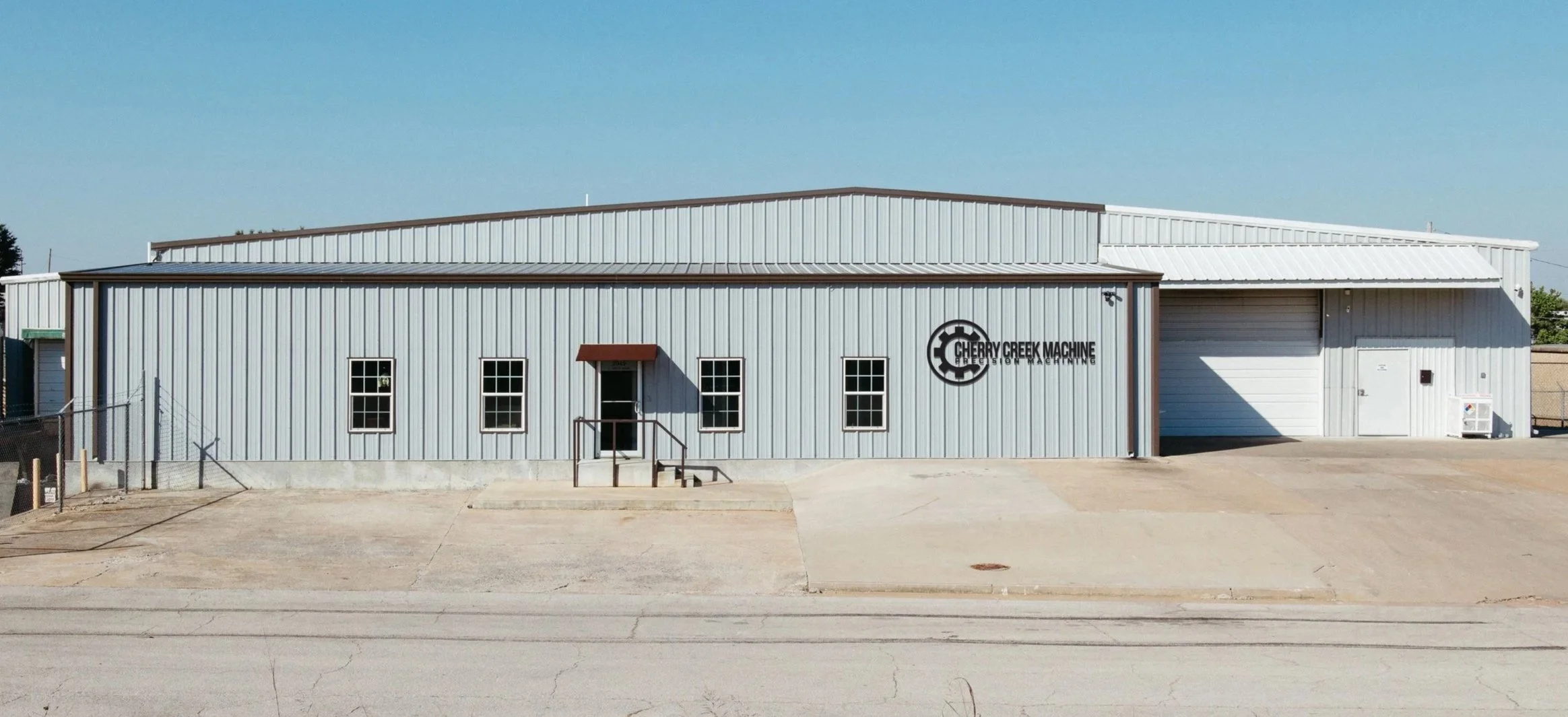 Front view of a metal industrial building with the sign 'Cherry Creek Machine' on the wall, three windows, a door with a small staircase, and a large garage door.