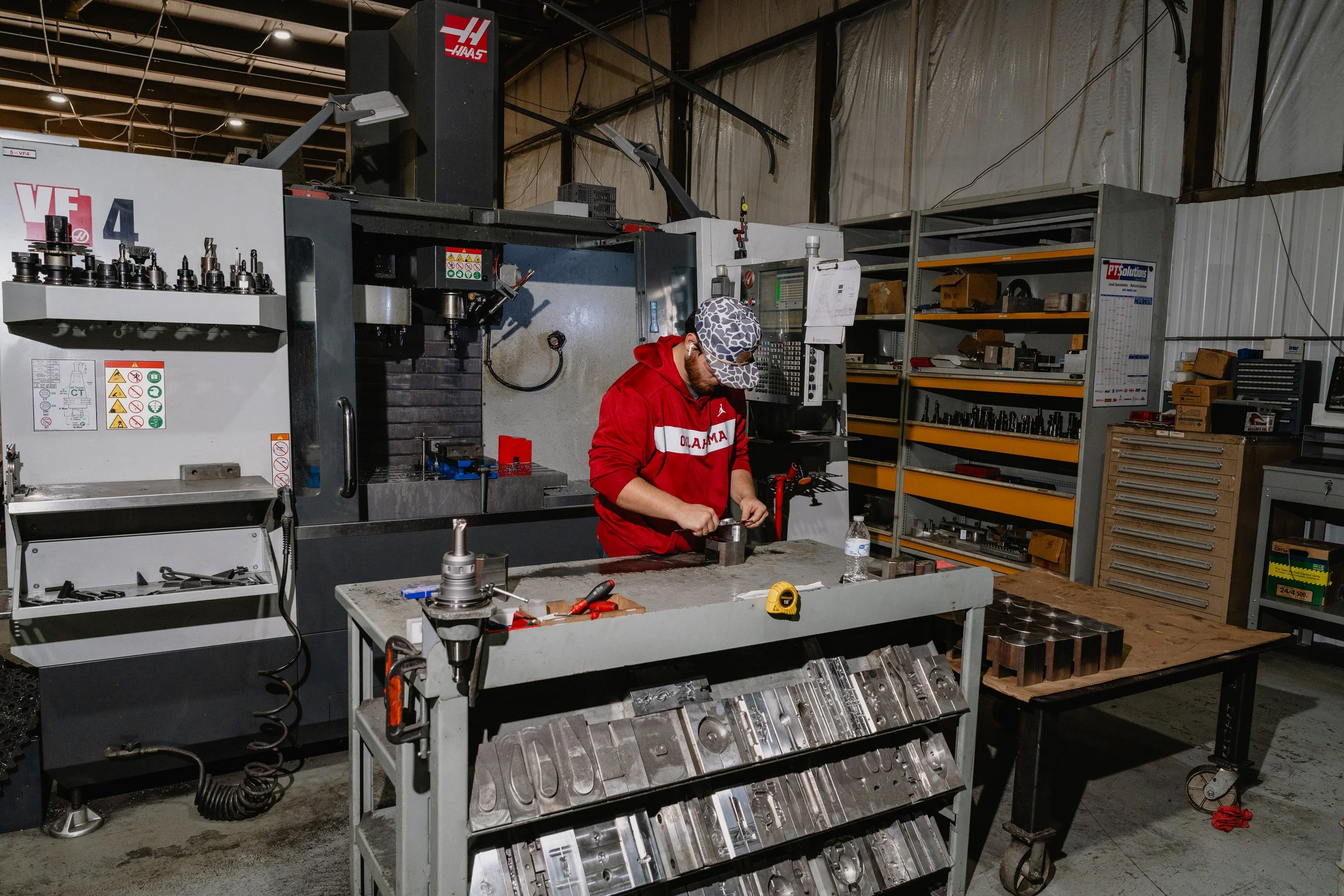 A man in a red Oklahoma hoodie and a camouflage baseball cap working with tools at a metalworking station in a workshop, surrounded by machinery and shelves with various machine parts.