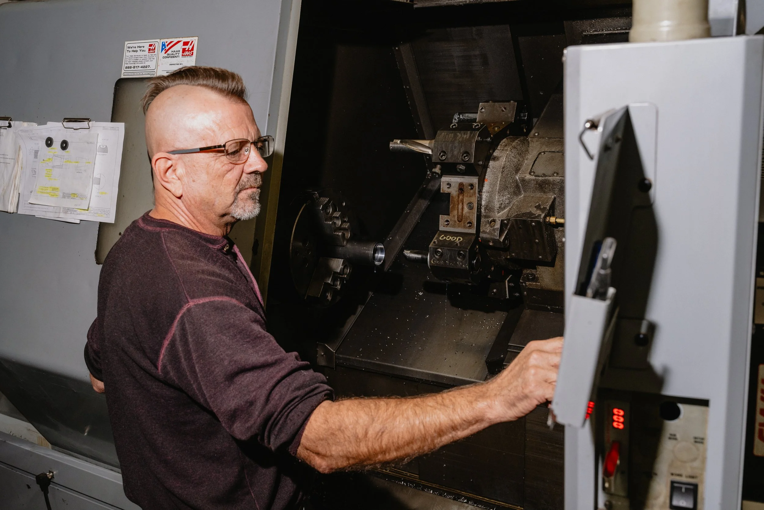 A man with glasses and a crew cut hairstyle working with a CNC machine, wearing a burgundy and black long sleeve shirt in a workshop.